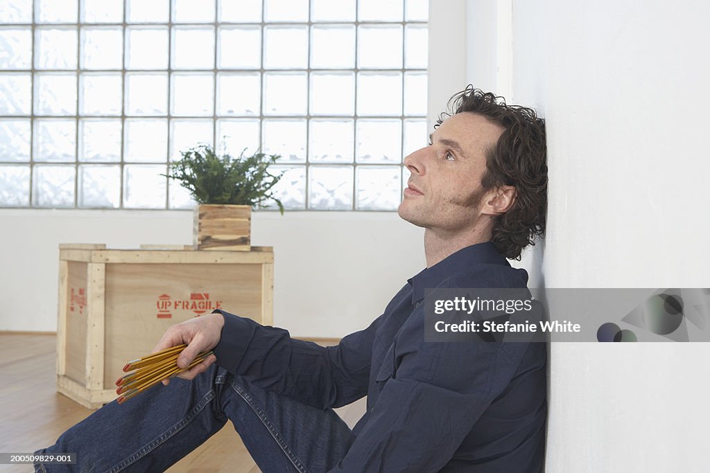 Man sitting on wooden floor, holding pencils, side view