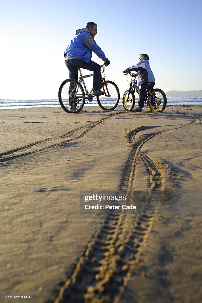 Father and son (6-8) riding bicycles on beach, tracks in foreground