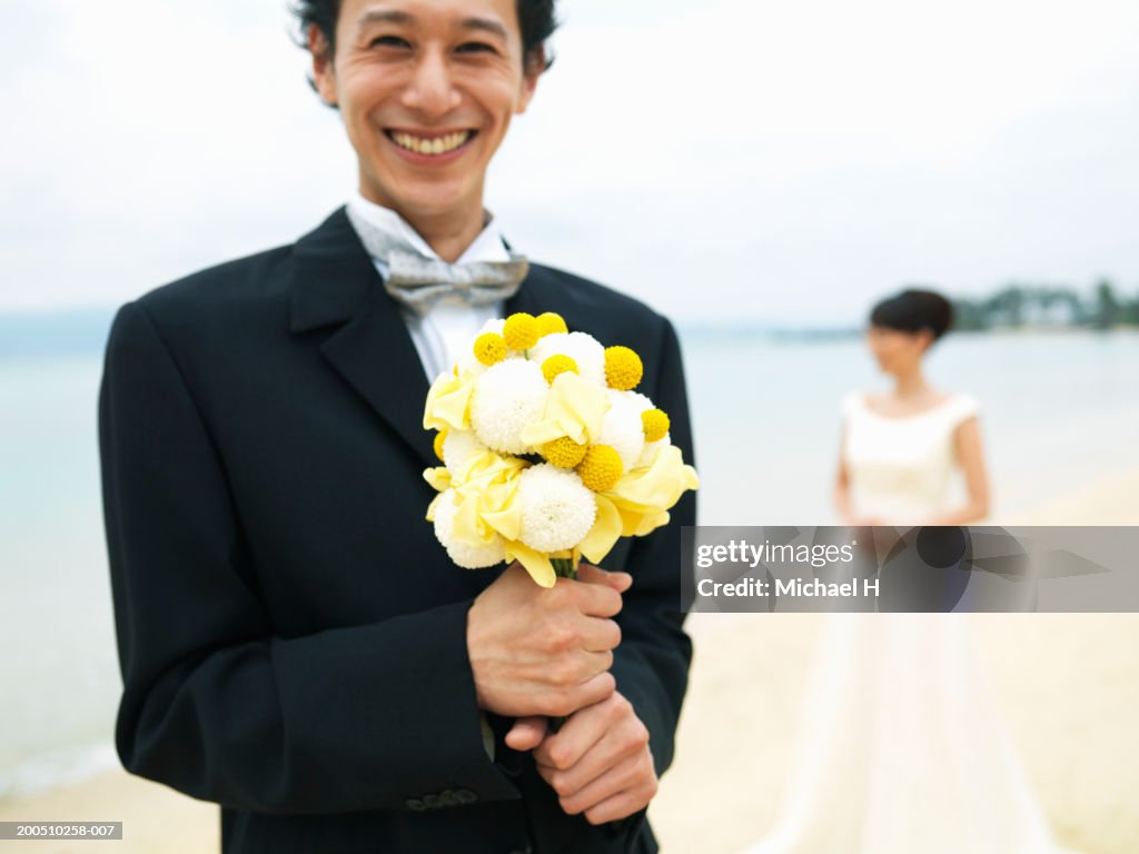 Groom holding bouquet of flowers on beach, with bride in background