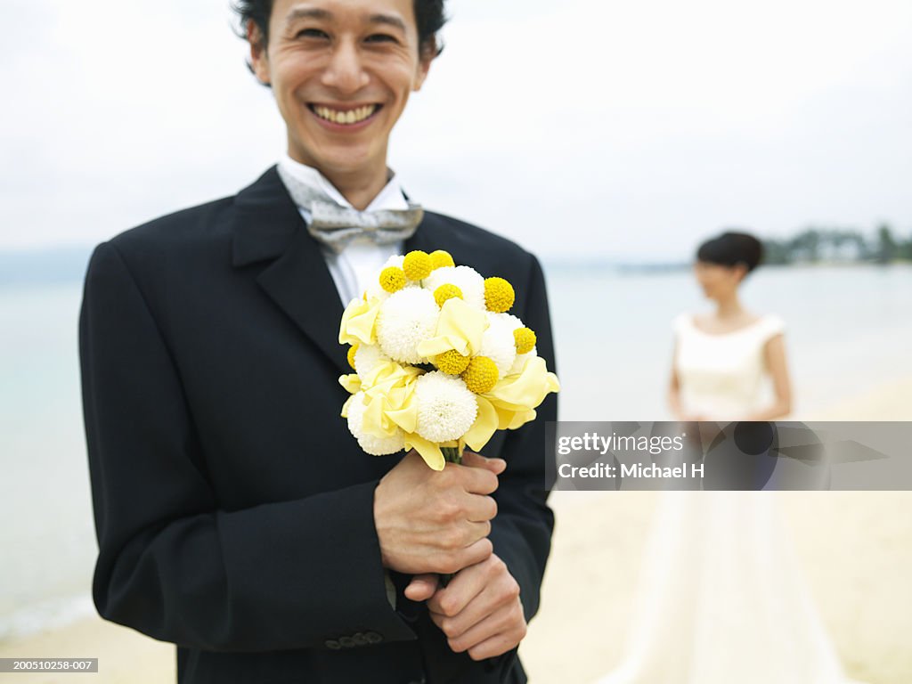 Groom holding bouquet of flowers on beach, with bride in background