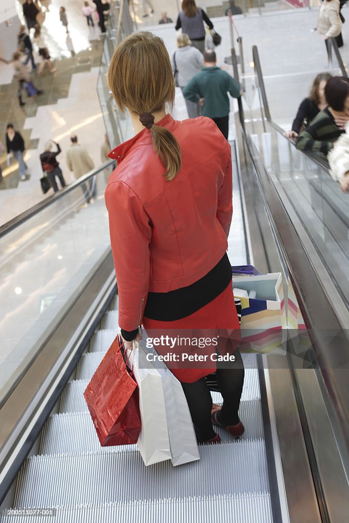 Young woman on escalator, holding shopping bags, rear view