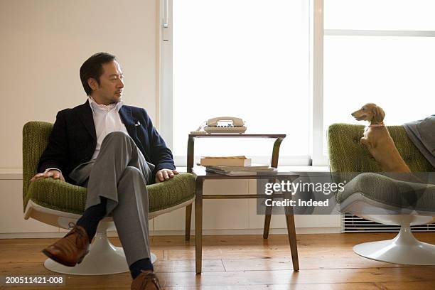 senior man relaxing in chair at home, looking at dog - asian man seated stockfoto's en -beelden