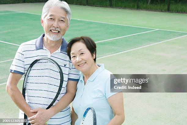 senior couple embracing, holding tennis racquets, smiling, portrait - japanese tennis photos et images de collection