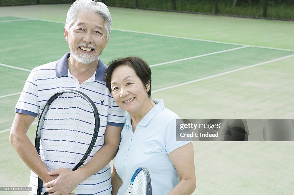 Senior couple embracing, holding tennis racquets, smiling, portrait