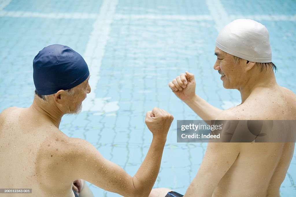 Two senior men sitting on edge of swimming pool, clenching fists