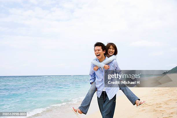 young man giving young woman piggyback ride on beach - japanese couple beach stock pictures, royalty-free photos & images