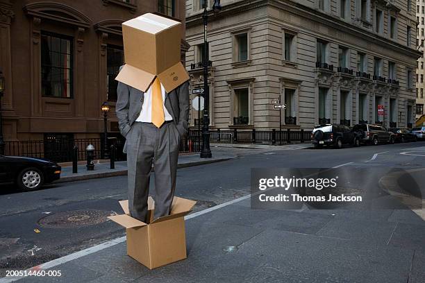 businessman standing in cardboard box and wearing one on head on city street - indifferenza foto e immagini stock