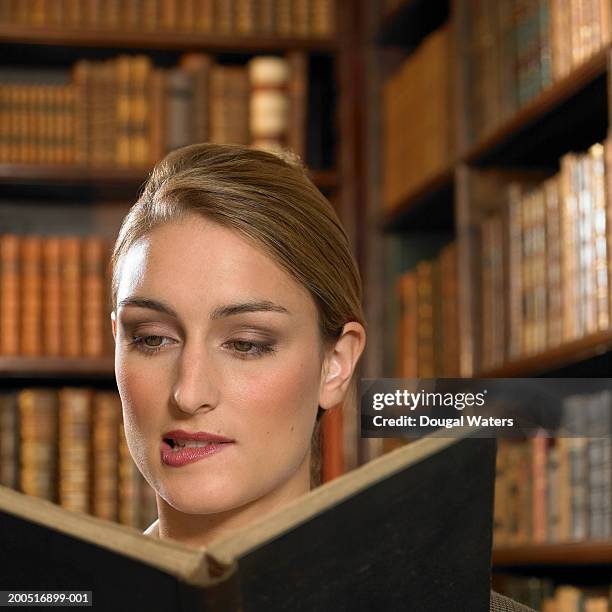 young woman biting lip as she reads book in library - hardbound stock pictures, royalty-free photos & images