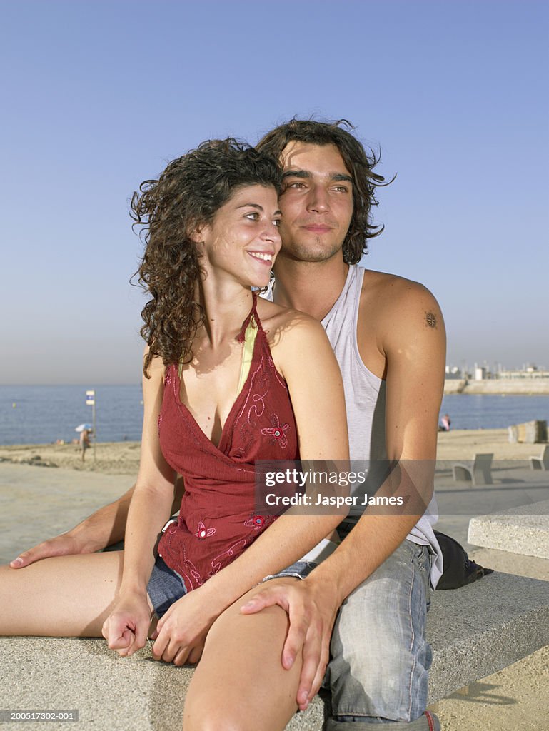 Young couple sitting on bench on beach promenade, smiling