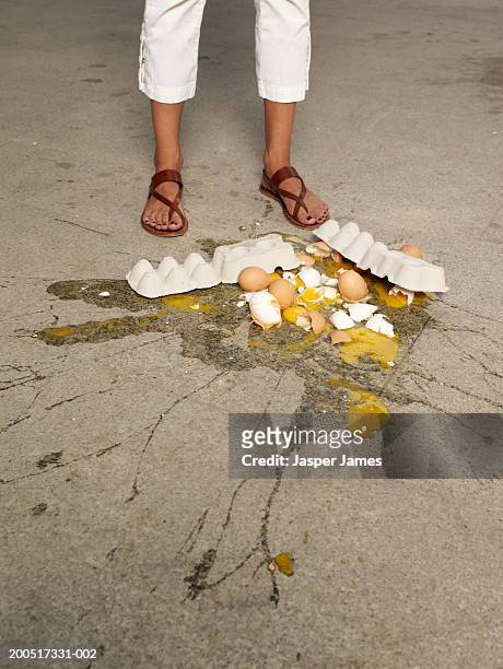woman standing beside carton of broken eggs on pavement, low-section - broken heel stock-fotos und bilder