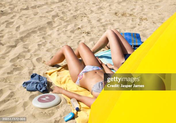 two young women lying on beach, obscured by yellow sunshade - sunbathing stock pictures, royalty-free photos & images