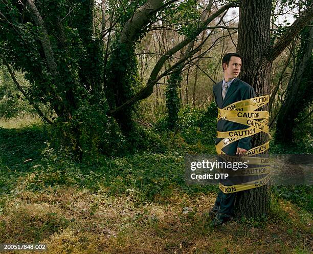 businessman tied to tree with yellow 'caution' tape in scrubland - amarrado fotografías e imágenes de stock