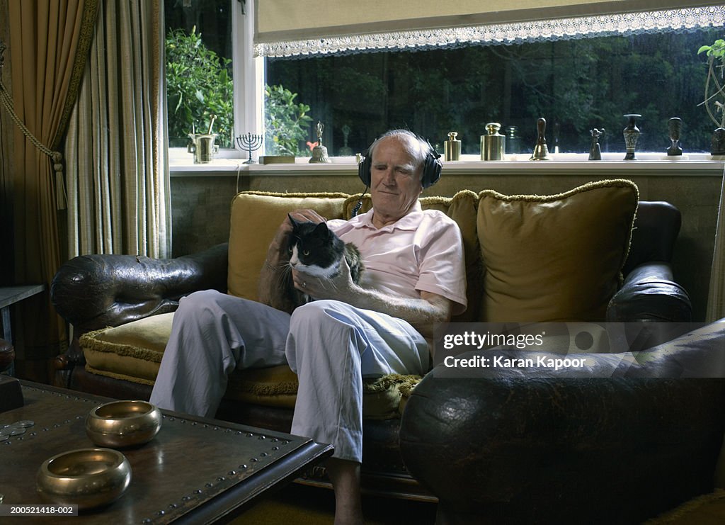 Senior man sitting on sofa, holding cat, wearing headphones