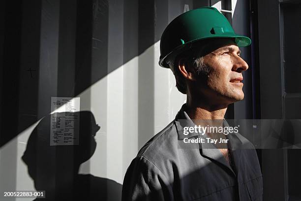 man standing beside cargo container, wearing hard hat, side view - blue collar portrait stock-fotos und bilder