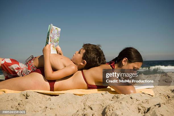 boy (4-6) lying on mother's back at beach, both reading books - beach book reading stock pictures, royalty-free photos & images