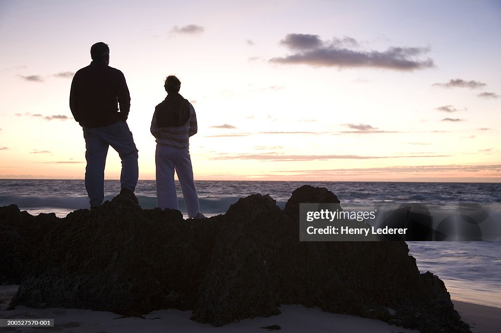 Silhouette of mature couple standing on rock, looking at sea, rear view