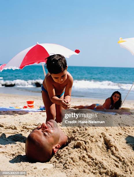 boy (3-5) burying father in sand at beach - bury fotografías e imágenes de stock