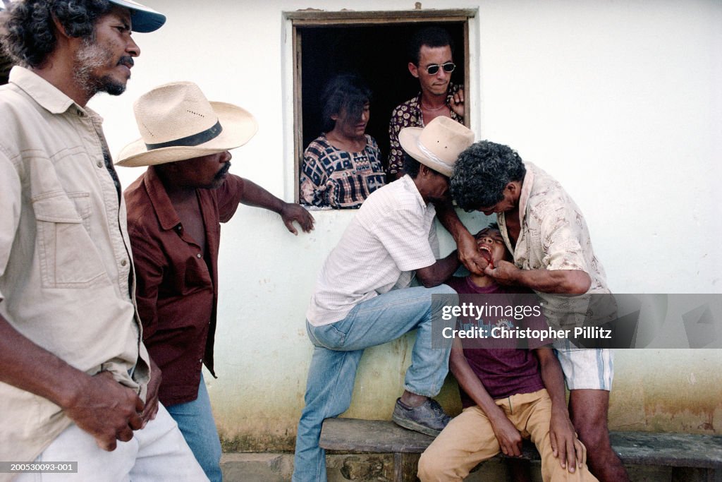 Brazil, Bahia, boy having teeth tended to by two men outside house