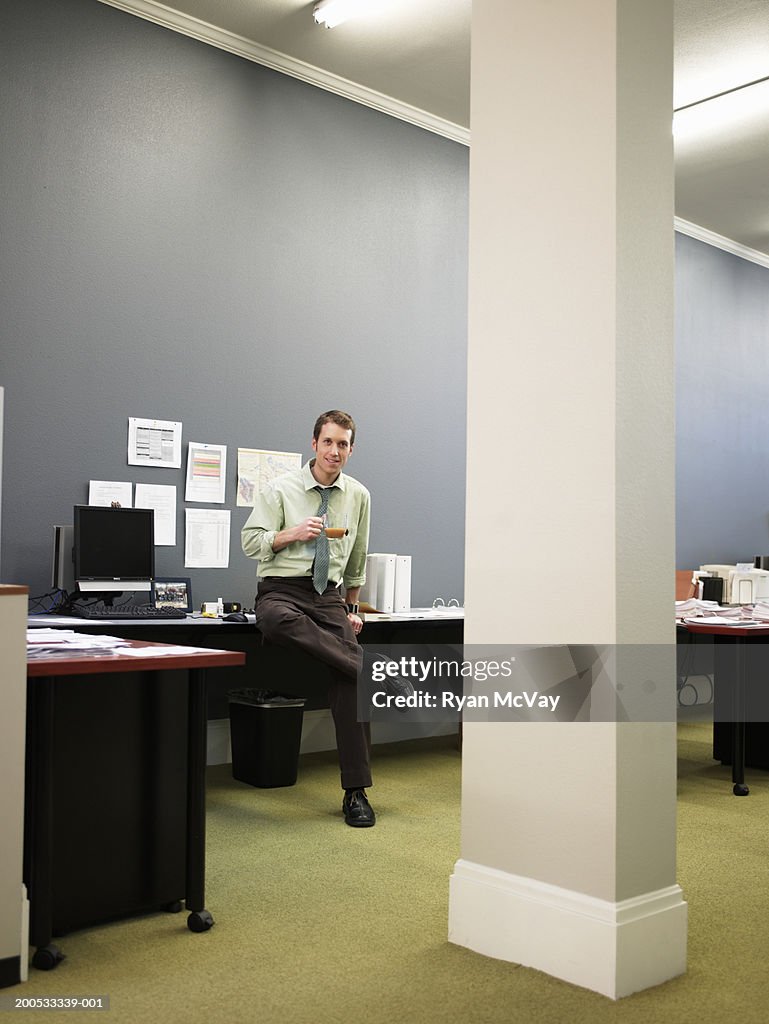 Young businessman sitting atop desk in office, portrait