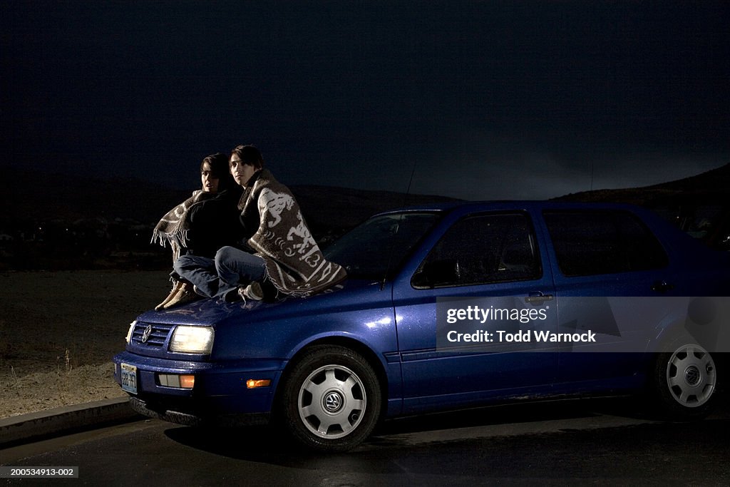 Young couple sitting on car wrapped in blanket, night