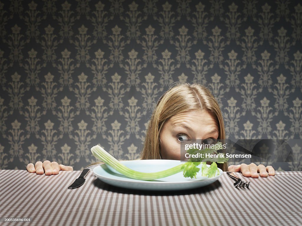 Young woman peering over dinner table, looking at celery on plate