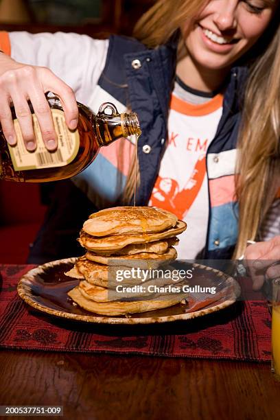 young woman pouring maple syrup on stack of pancakes, smiling - maple syrup pancakes fotografías e imágenes de stock