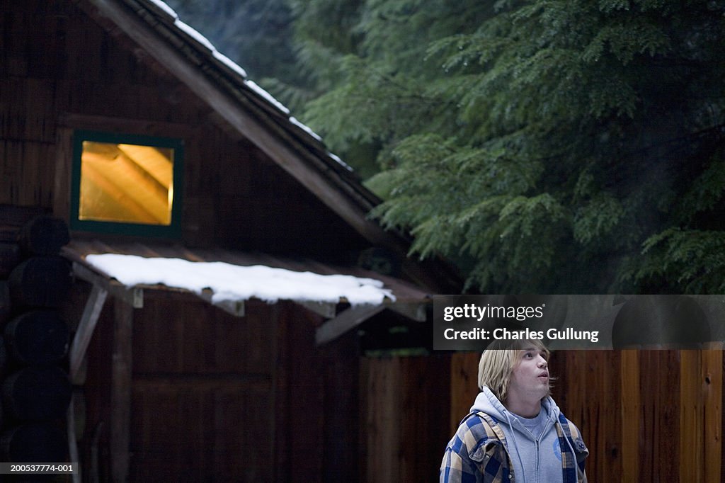 Teenage boy (16-18) standing outside log cabin, looking up