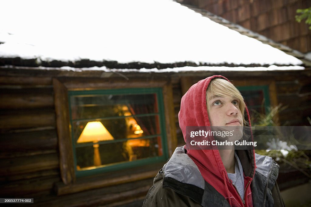 Teenage boy (16-18) standing outside log cabin, looking up