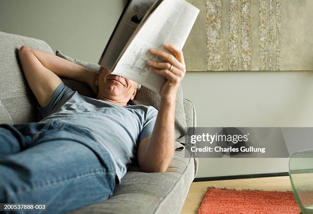 mature man reading magazine on sofa, smiling, hand behind head - reading magazine stock-fotos und bilder