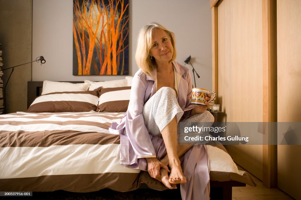 Mature woman with coffee mug sitting on edge of bed, portrait