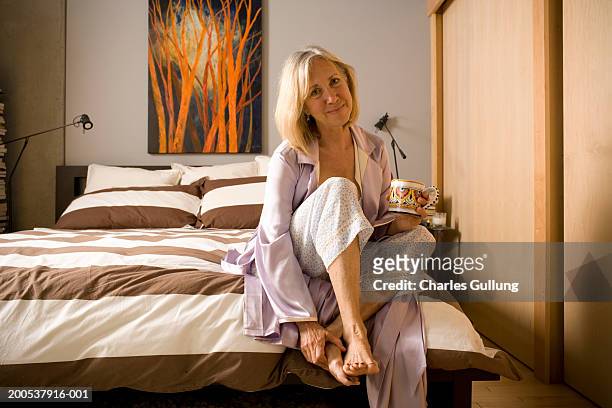 mature woman with coffee mug sitting on edge of bed, portrait - older woman legs - fotografias e filmes do acervo