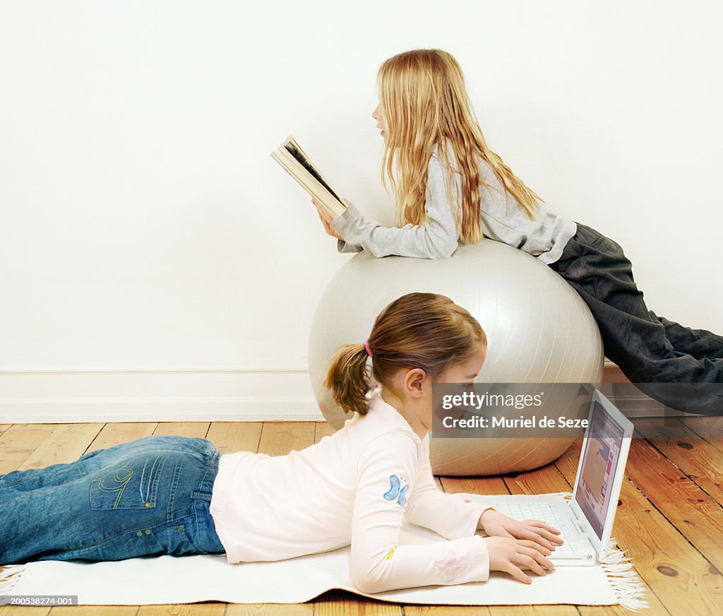 Two girls (7-9) relaxing in living room, reading and looking at laptop