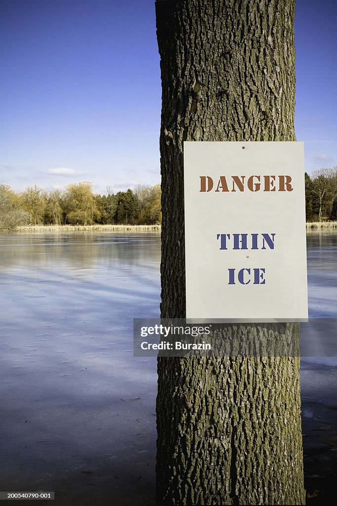 Thin ice warning sign on tree by lake, winter