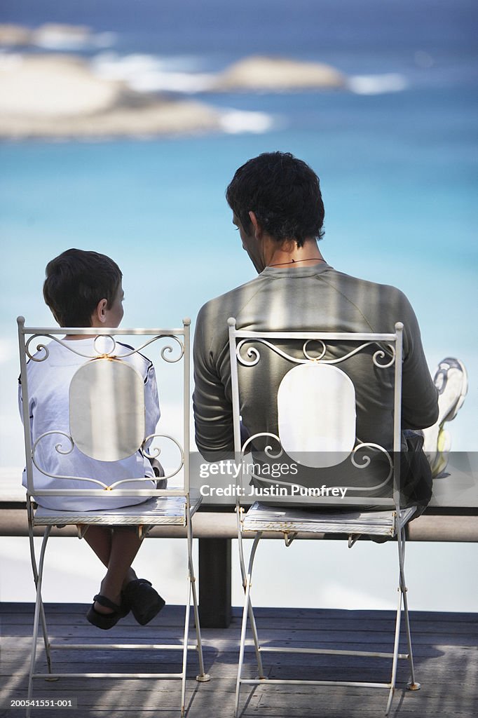 Father and son (6-8) sitting in outdoor chairs, rear view