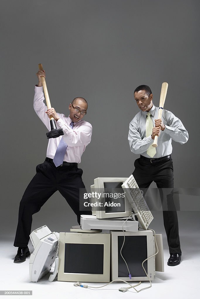 Two businessmen holding sledgehammer and baseball bat, near pile of old technology