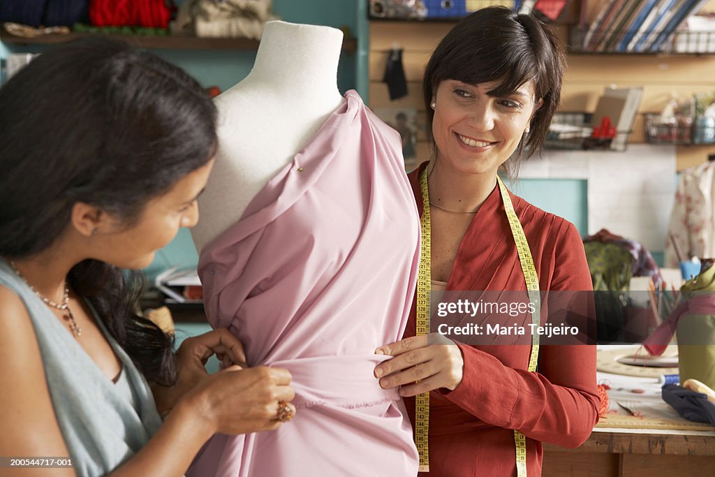 Two female fashion designers dressing mannequin in studio, smiling