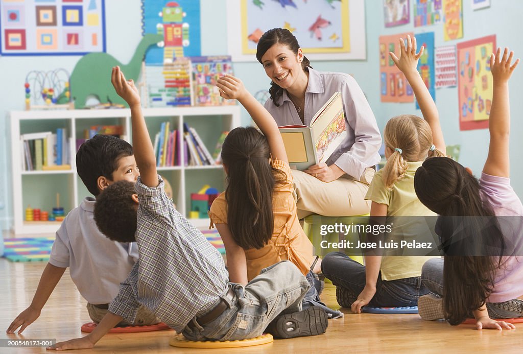 Teacher reading book in classroom, children (2-7) raising hands