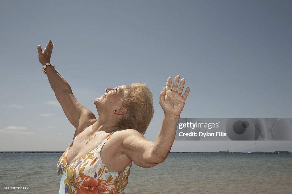 Senior woman wearing swimming costume on beach, arms raised, smiling