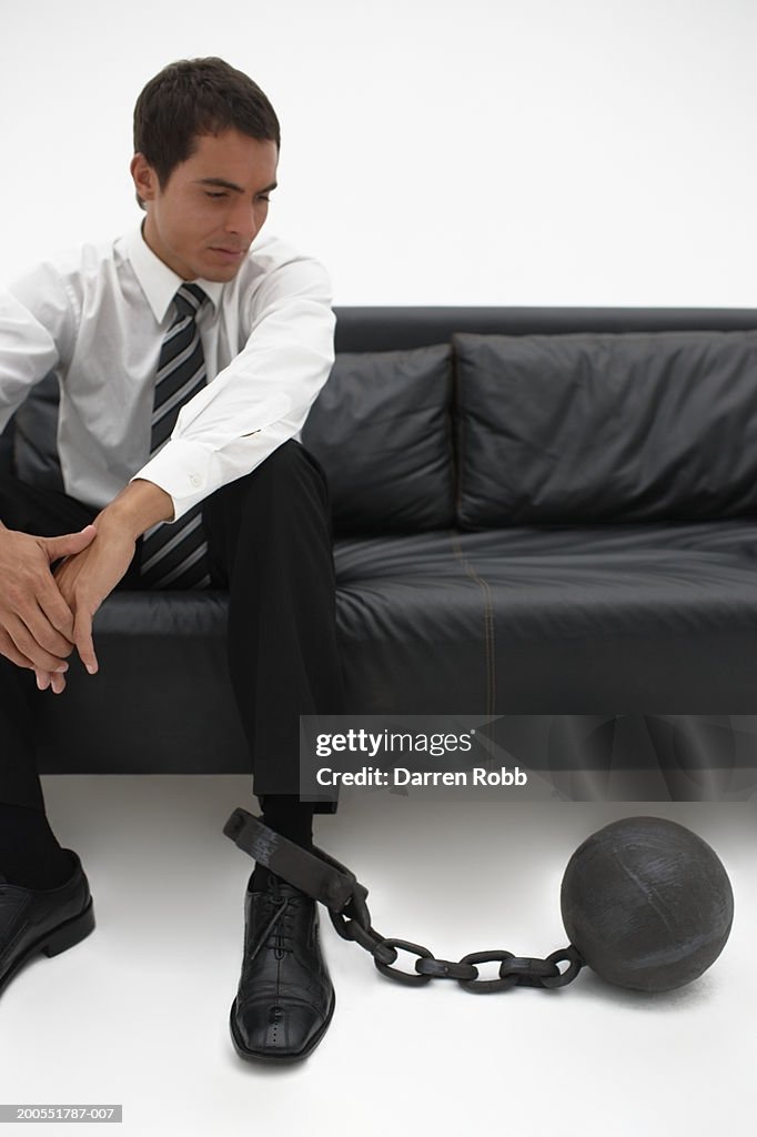 Young businessman sitting on sofa looking at ball and chain attached on leg