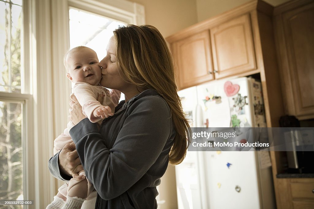 Mother kissing baby boy (15-18 months) in kitchen, side view