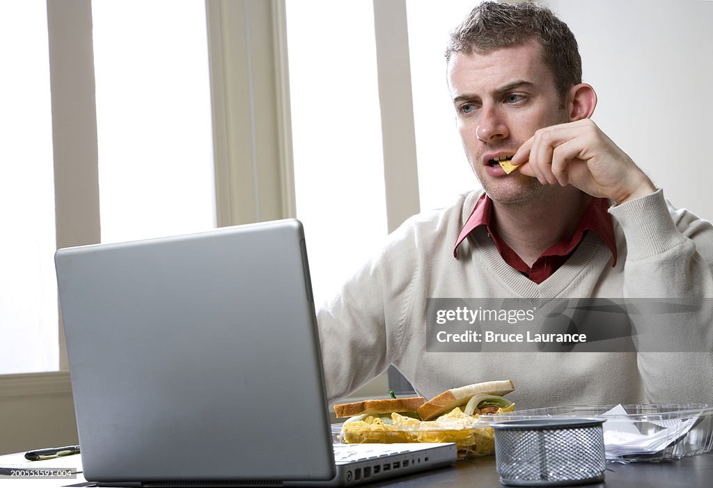 Young man sitting at desk, using laptop, having lunch