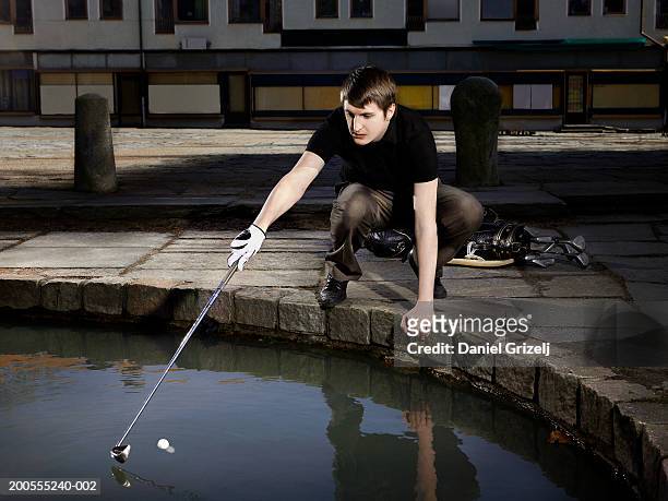 young man playing city golf, trying to take ball out of pond using golf club - golf short iron stock pictures, royalty-free photos & images