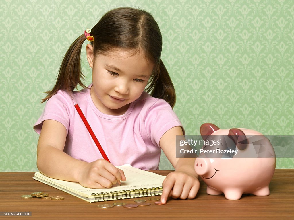 Girl (6-7) at desk, counting and recording contents of piggy bank