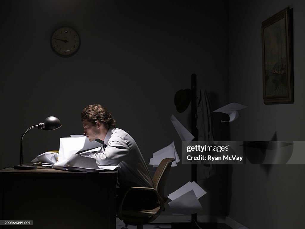 Young office worker sitting at desk, papers flying behind, side view
