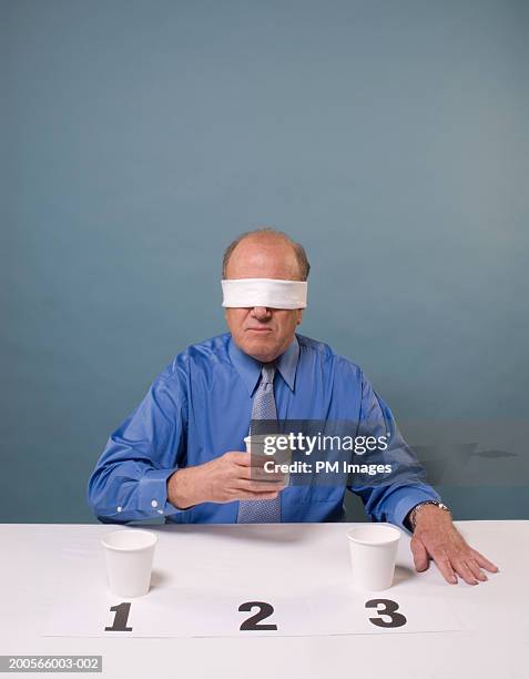 blind folded middle aged man taking taste test, holding one of three cups - blindfold stock pictures, royalty-free photos & images