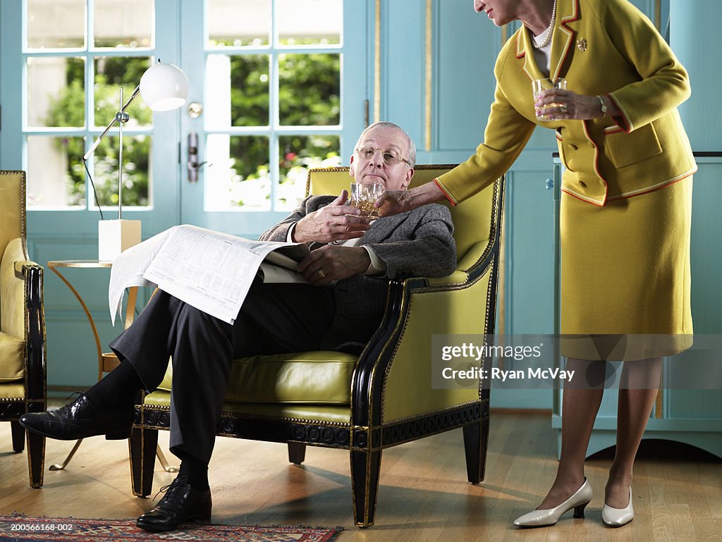 Senior woman handing drink to senior man sitting in armchair