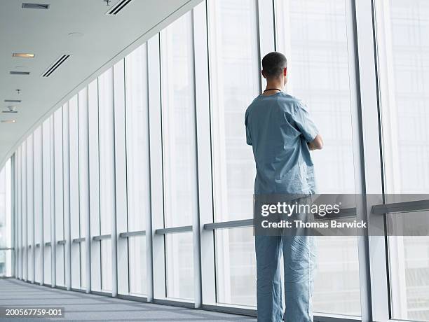 man in scrubs looking out window of hospital corridor - medizinerkleidung stock-fotos und bilder