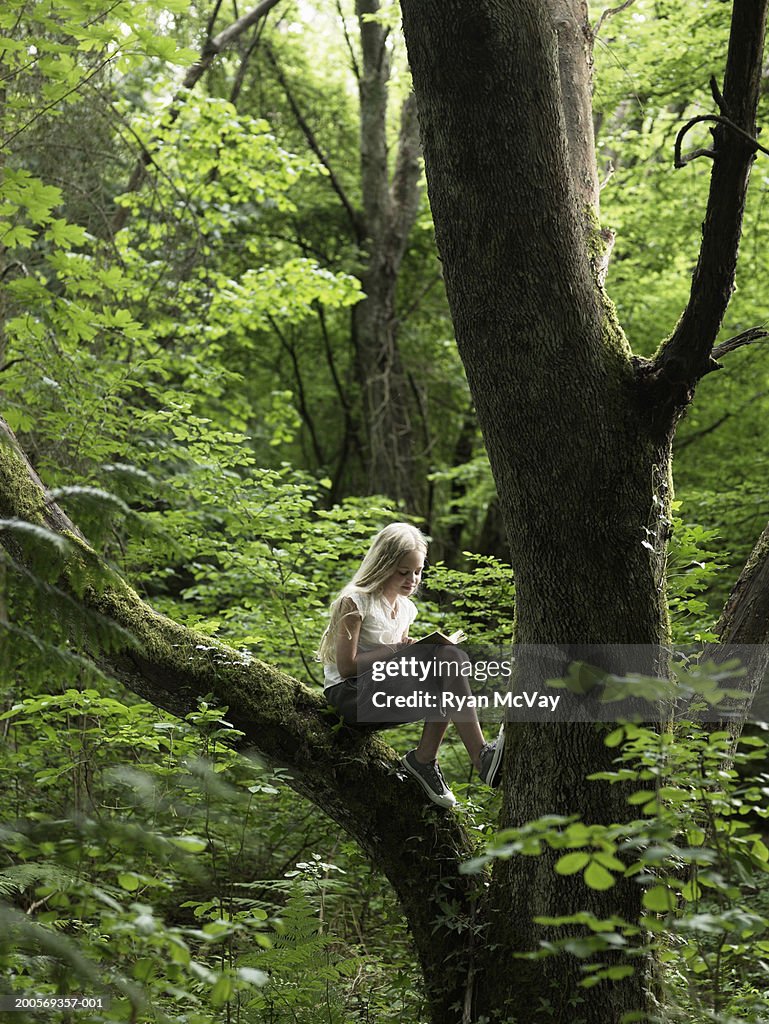 Young girl (10-11) climbing tree in forest, reading book