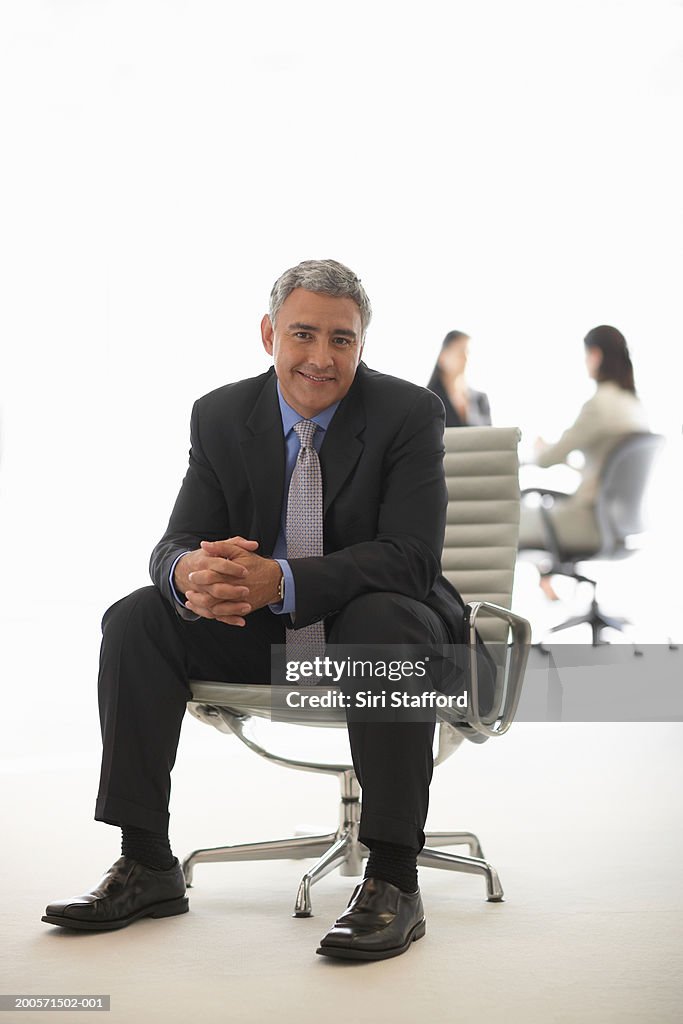 Mature businessman sitting in office chair, women in background