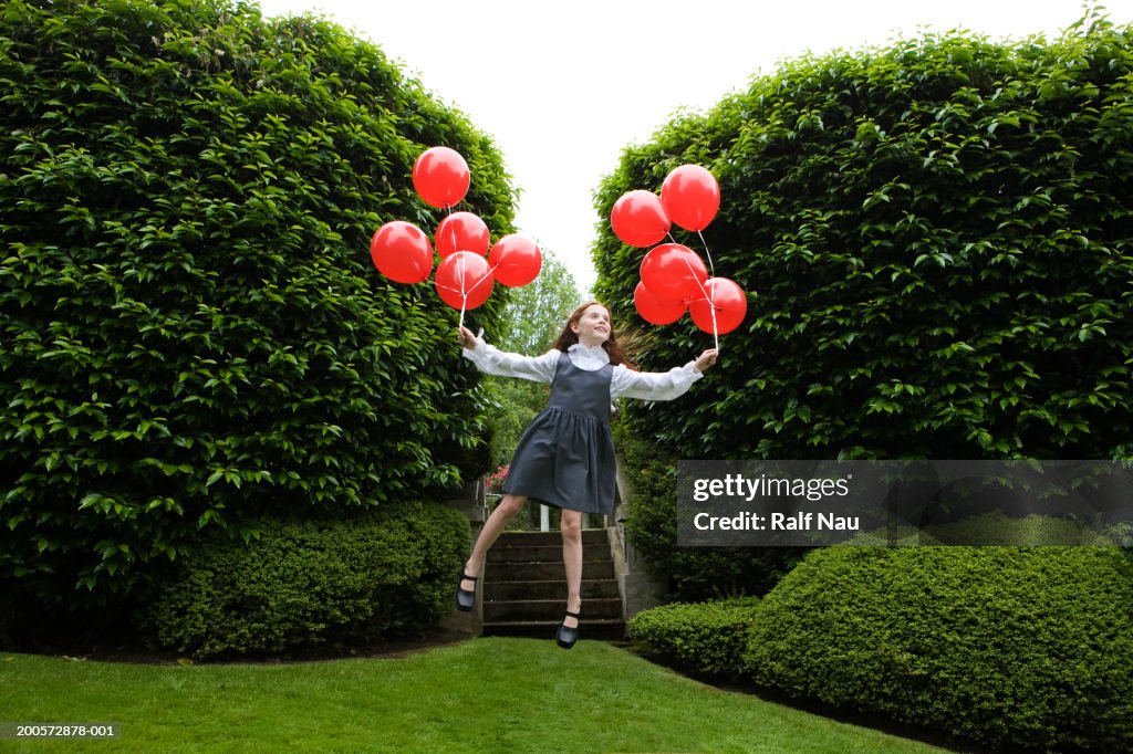 Girl (5-7) holding red balloons, floating in garden